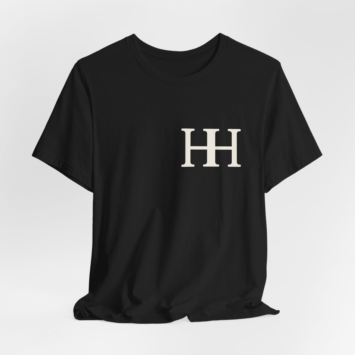 Mockup: black Soft tee on a hanger against a light wall, ivory HH mark on the chest