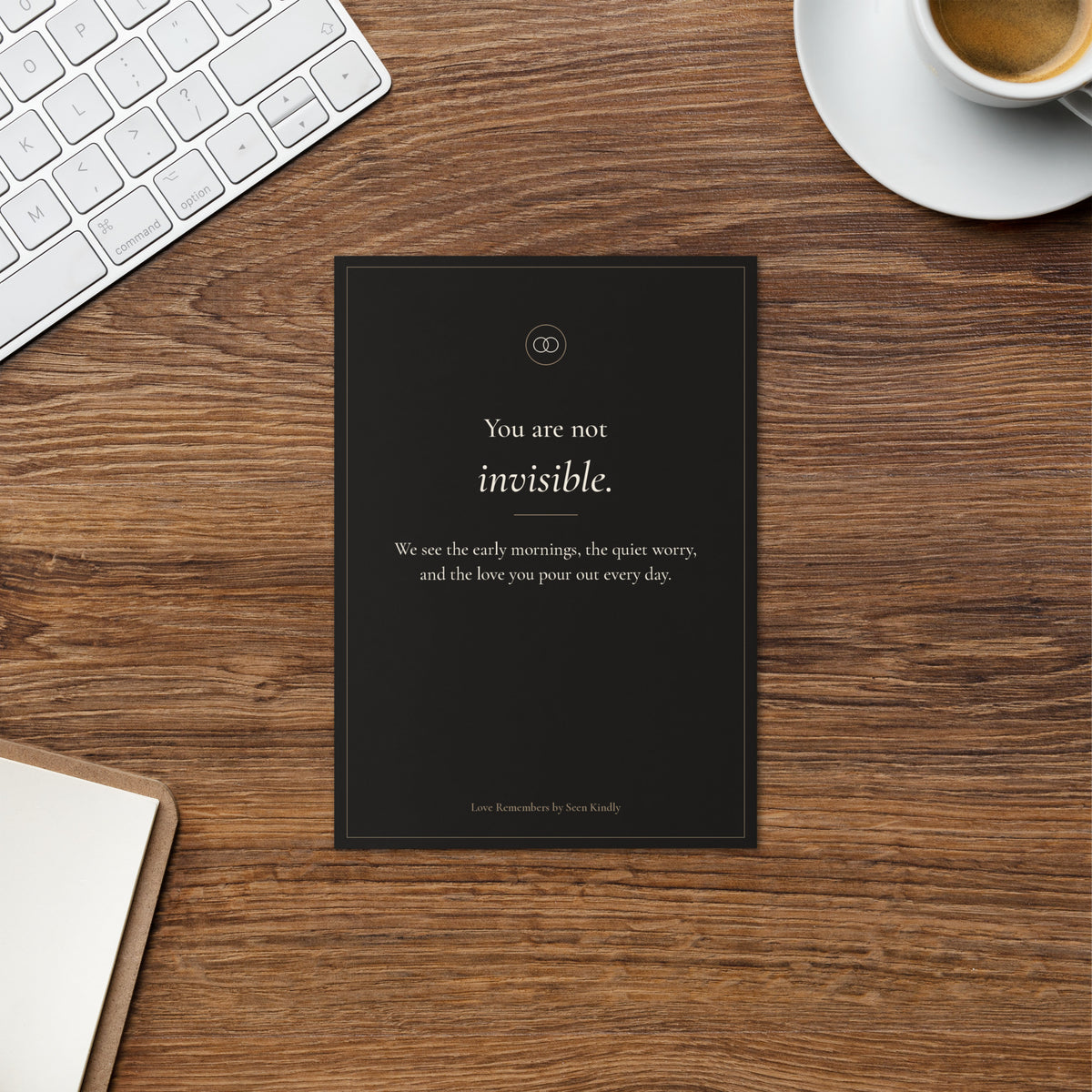 A black Love Remembers card reading You are not invisible, on a warm wood desk beside a cup of coffee, the kind of card tucked into a Haven Box.