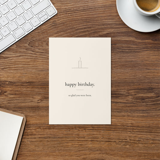 Happy Birthday card on a warm wood desk beside a cup of coffee, with a single candle and the words happy birthday. so glad you were born.